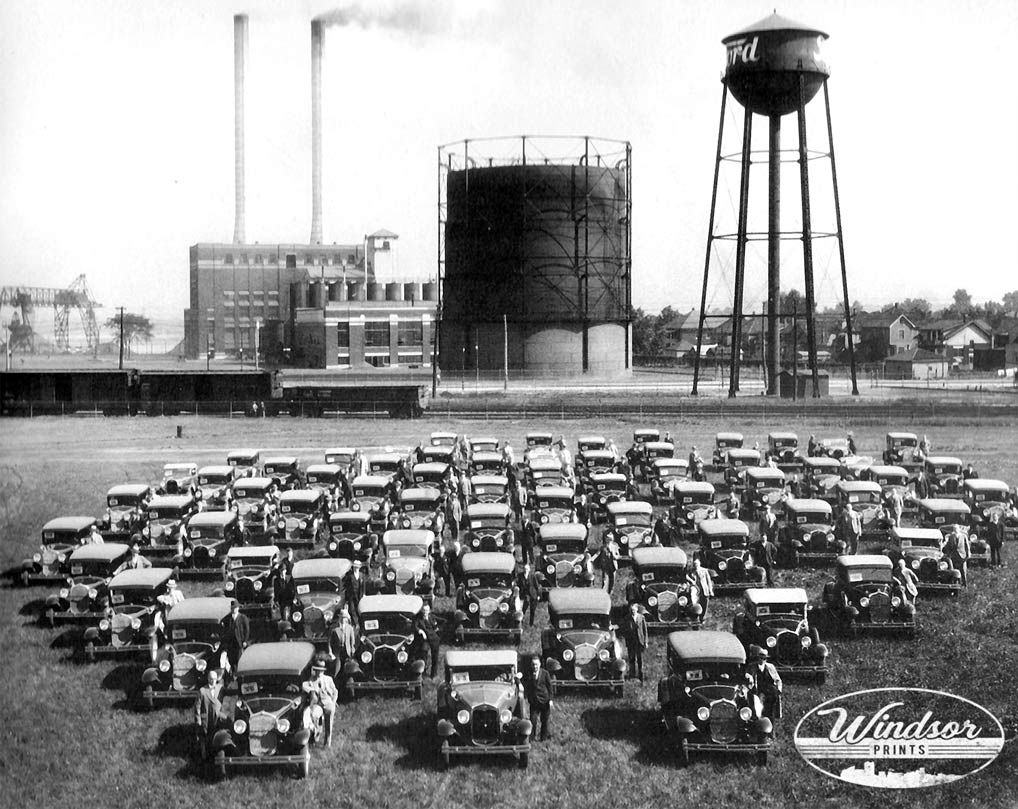Historic photo of dozens of Ford cars lined up in a field at the Ford plant in Ford City, Windsor, Ontario, with smokestacks and the Ford water tower behind